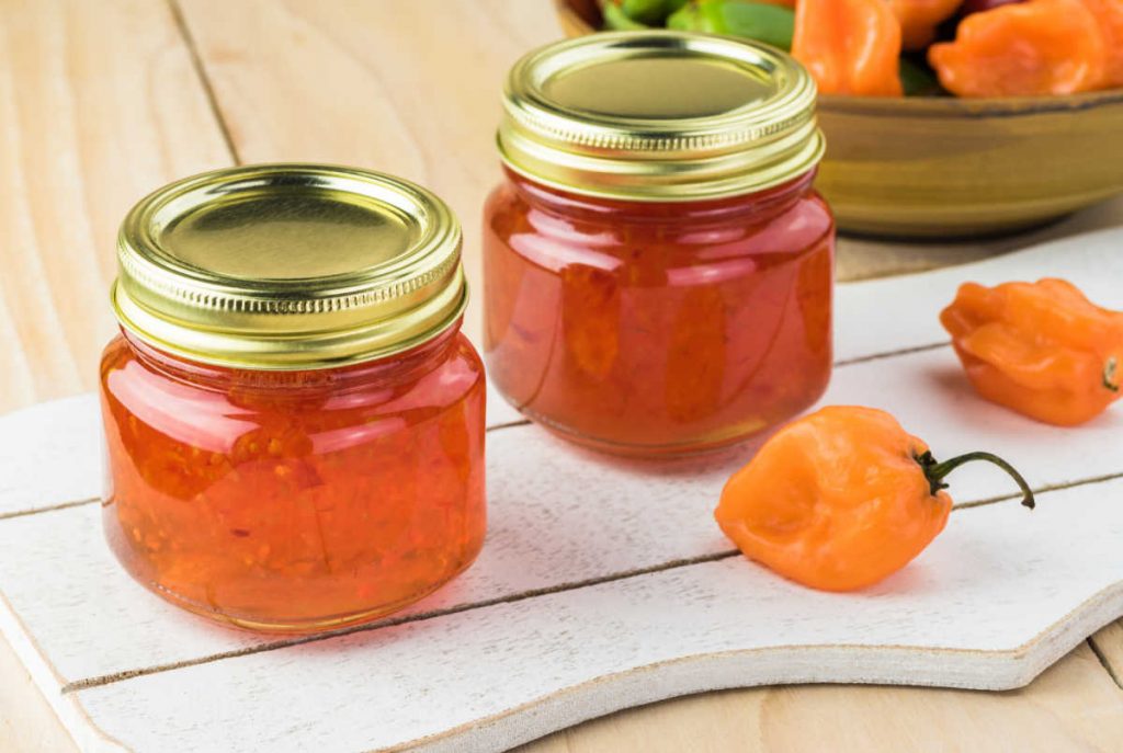 Two small jars of pepper jelly on a cutting board with orange habanero peppers around it.